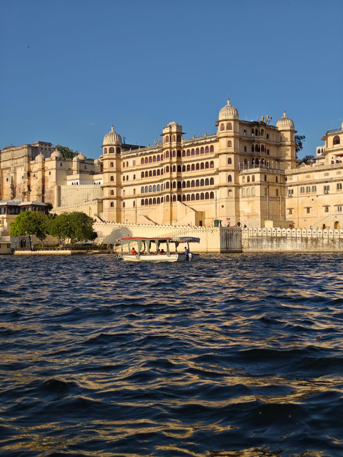 Scenic view of the historic City Palace in Udaipur, India, with a boat on Lake Pichola at sunset.